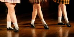 Three dancers in plaid skirts performing on a wooden stage