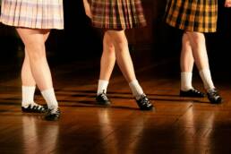 Three dancers in plaid skirts performing on a wooden stage
