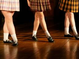 Three dancers in plaid skirts performing on a wooden stage